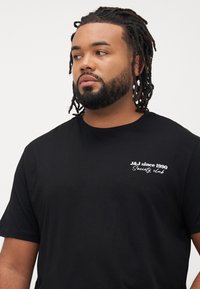Zwarte katoenen T-shirt met een ronde hals, korte mouwen en witte tekst op de borst die leest: "J&J sinds 1990 Society Club."