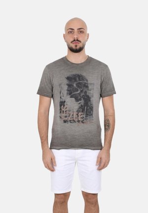 Uomo con barba che indossa una t-shirt grafica grigia e pantaloncini bianchi, in piedi davanti a uno sfondo neutro, con espressione facciale neutra.