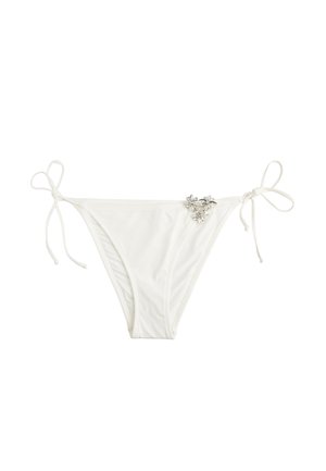 Weiße Bikinihöschen mit seitlichen Bindebändern und einem kleinen Cluster aus silbernen, blumenförmigen Verzierungen auf der vorderen rechten Seite.