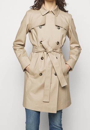 Femme portant un trench-coat beige croisé avec une ceinture à la taille et des poches latérales, associé à un jean bleu.