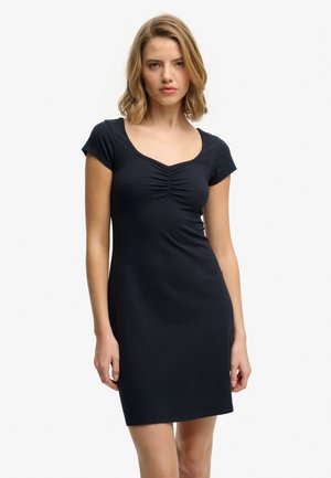 Vrouw met schouderlang blond haar, gekleed in een nauwsluitende zwarte jurk met korte mouwen en een geplooide halslijn, staand tegen een effen witte achtergrond.