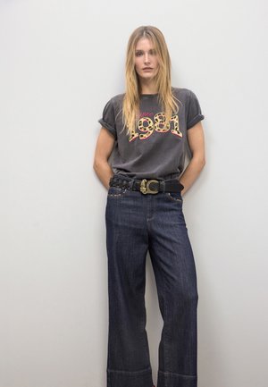 Camiseta gris con "1981" en estampado de leopardo, combinada con vaqueros de pierna ancha azul oscuro. Incluye un cinturón negro trenzado con detalle de hebilla dorada.