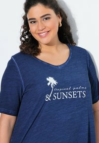 T-paita lyhyillä hihoilla tummansinisena, V-pääntiellä, jossa on valkoista tekstiä ja palmupuugrafiikka. Pehmeä tuntuma, rento malli ja mukava muotoilu.
