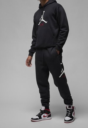 Osoba ubrana w czarną bluzę z kapturem i spodnie dresowe z białymi i czerwonymi logo Jumpman, w zestawieniu z czarnymi, białymi i czerwonymi wysokimi sneakersami.