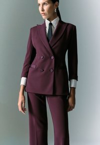 Abito doppiopetto in un profondo bordeaux, caratterizzato da un design strutturato, rever appuntiti e polsini bianchi a contrasto. Taglio sartoriale con una silhouette moderna.