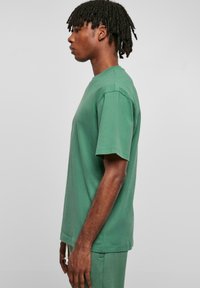 Tricou din bumbac verde cu un croi lejer, mâneci scurte și decolteu rotund. Textură netedă, cu cusături minime și fără grafică vizibilă.