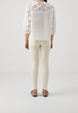 Personne portant un chemisier blanc à œillets avec découpes florales et pantalon slim crème, vue de dos, debout sur un sol blanc.