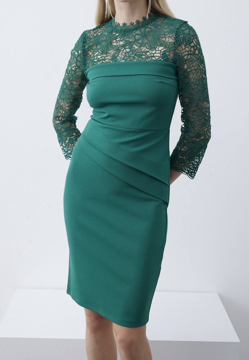 Femme portant une robe verte ajustée avec des manches en dentelle florale et le haut de la poitrine, debout devant un fond clair uni.