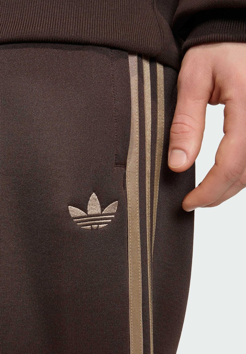 Hand ruht neben dunkelbrauner Adidas Trainingshose mit beigen drei Streifen und dem Trefoil-Logo am Oberschenkel.