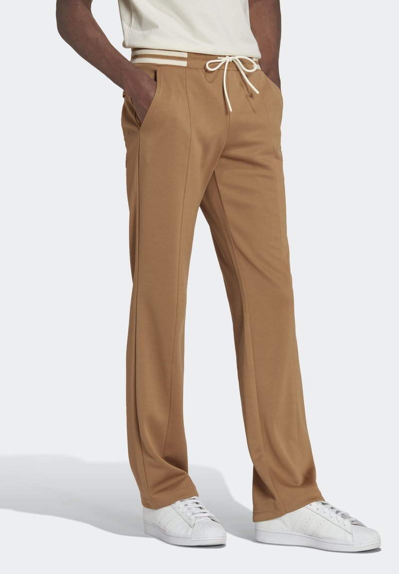 Bruna, skräddarsydda sweatpants med dragsko i midjan, sidfickor och insvängda ben, i kombination med vita sneakers på en grå yta.