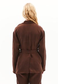 Hnědá sako s páskem, hladkým povrchem, dlouhými rukávy a klopovým límcem. Má zadní rozparek pro snadný pohyb.