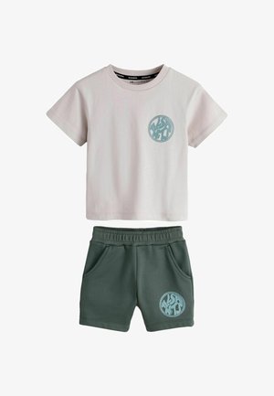Beige top med korte ærmer og grønne shorts med elastik i taljen, begge med et rundt blåt logo på venstre side.