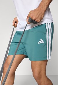 Zöld sportnadrág fehér oldalcsíkokkal és hímzett logóval. Az anyag könnyűnek és simának tűnik, alkalmas sporttevékenységekhez.