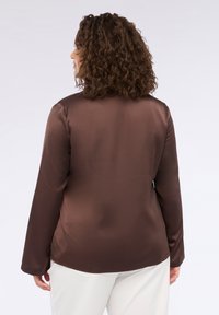 Blusa di raso marrone con maniche lunghe, texture liscia e spacchi laterali; indossata con pantaloni di colore chiaro, vista da dietro.