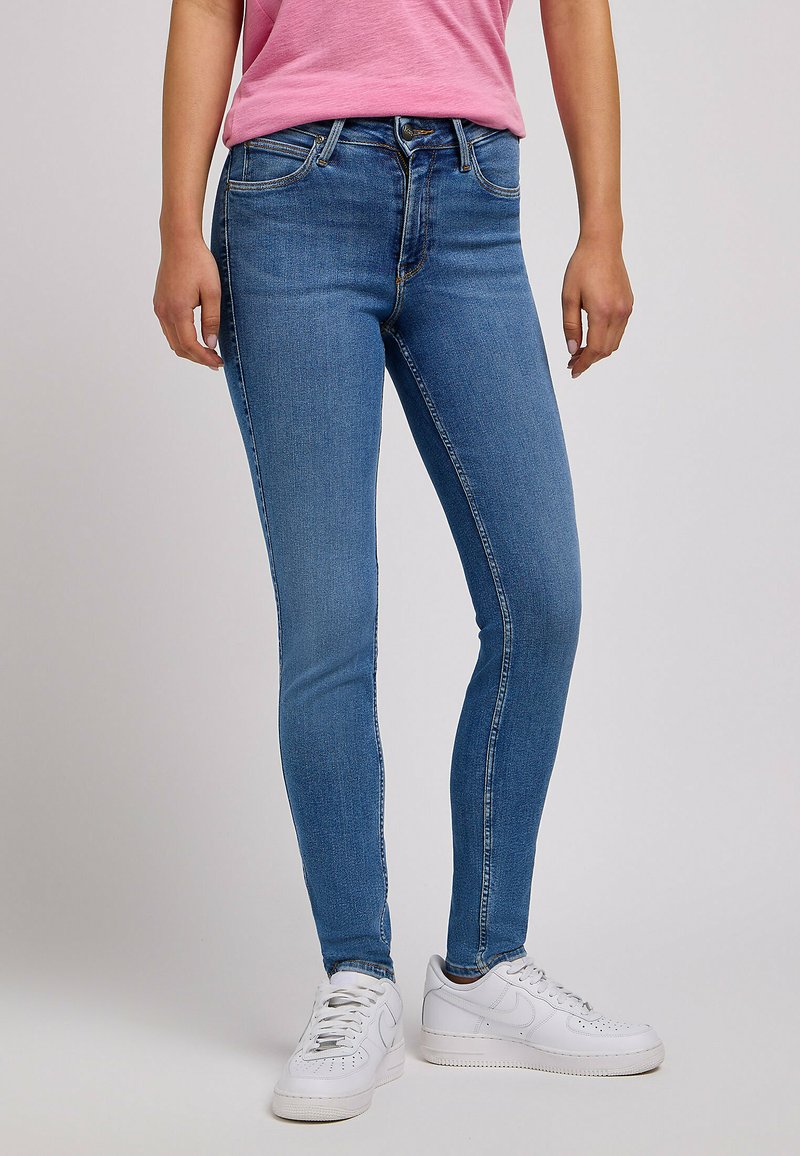 Cienkie niebieskie dżinsy z pięcioma kieszeniami, złotymi przeszyciami i paskiem ze szlufkami. W połączeniu z białymi trampkami.