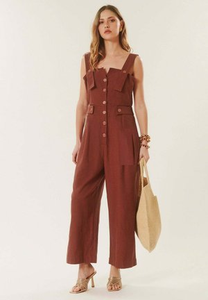 Femme debout portant une combinaison sans manches couleur rouille, boutonnée et munie de poches, tenant un sac cabas tissé beige, et portant des sandales à talons.