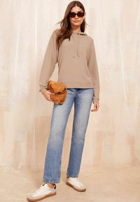 Felpa beige con cappuccio e orlo a costine, jeans svasati blu e sneakers bianche con dettagli marroni. Il modello tiene una borsa marrone.