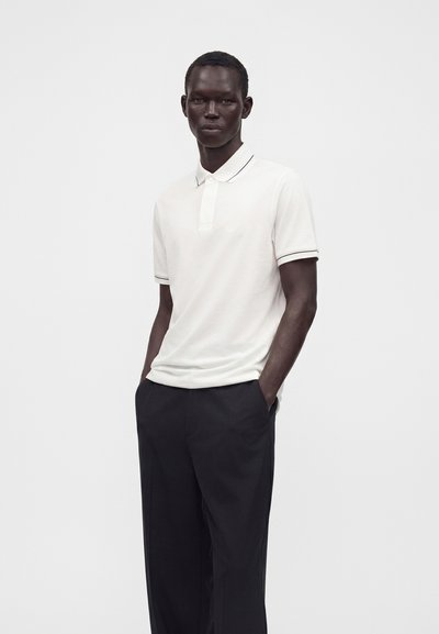 Homme à la peau foncée portant un polo blanc et un pantalon noir, debout les mains dans les poches devant un fond uni.