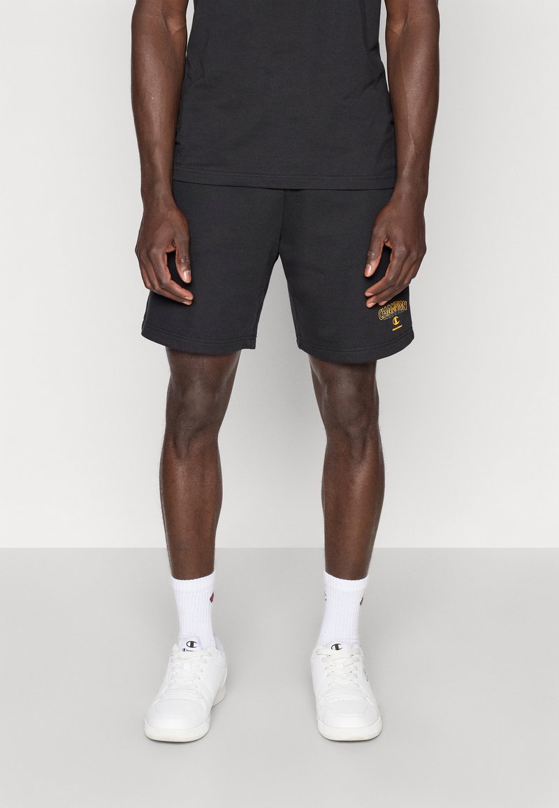 Μαύρα αθλητικά σορτς από βαμβάκι με ευθεία κοπή, που φέρουν ένα μικρό κίτρινο λογότυπο στο αριστερό πόδι και συνδυάζονται με λευκές κάλτσες και αθλητικά παπούτσια.