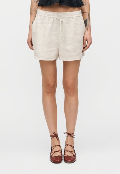 Shorts en lin beige clair avec une taille élastique et un cordon de serrage. Portés avec des chaussures plates marron à lacets, mettant en valeur une texture lisse et une coupe décontractée.