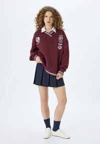 Bordeaux sweatshirt med vita ränder och tryckta detaljer, tillsammans med en marinblå plisserad kjol. Vita sneakers och marinblå strumpor fulländar outfiten.