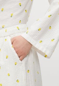 White textured fabric with small yellow lemon prints; features long sleeves and an elastic waist with pockets.