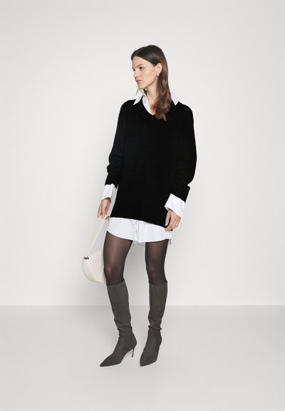 Schwarzer oversized Pullover über einem weißen Hemd, kombiniert mit grauen Wildleder-Stiefeln bis zum Knie und einer cremfärbigen Umhängetasche.