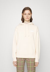 Sweat à capuche couleur crème avec une poche kangourou et un logo "Levi's" blanc. Dispose de cordons de serrage et de poignets côtelés, associé à un pantalon à carreaux.