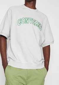 Jasnoszary sweatshirt z zielonym haftowanym napisem "CONVERSE", luźny krój, okrągły dekolt i krótkie rękawy, w połączeniu z zielonymi spodniami.