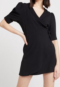 Black wrap dress with short puff sleeves, deep V-neckline, tied side accent, and smooth jersey texture. Versatile and fitted silhouette.