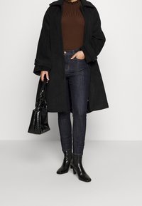 Manteau en mélange de laine noire avec boutons, pull brun côtelé, jean en denim foncé et bottes à talons noires. Tient un sac à main noir texturé.