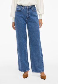 Φαρδιά μπλε τζιν παντελόνια με ψηλή μέση, διαθέτουν μπροστινές τσέπες και ξεθωριασμένη υφή, συνδυασμένα με καφέ ψηλές γόβες.