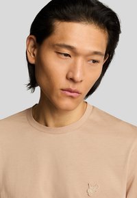 T-shirt beige a maniche corte con scollo rotondo e un piccolo logo ricamato a forma di farfalla sul petto. Tessuto di cotone morbido, texture liscia.