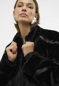 Czarna kurtka z sztucznego futra z wysokim kołnierzem, zapinana na zamek, o kontrastujących fakturach. Modelka pokazuje zaciśnięte pięści, nosząc srebrne kolczyki wyróżniające się.
