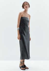 Trägerloses, knöchellanges Kleid aus strukturiertem, dunkelgrauem Stoff. Kombiniert mit schwarzen Sandalen mit überkreuzten Riemen und einem metallischen Armreif.