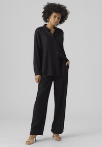 Camicia nera a maniche lunghe con bottoni abbinata a pantaloni neri a vestibilità ampia. Tessuto morbido con silhouette rilassata. Il modello indossa sandali con tacco beige.