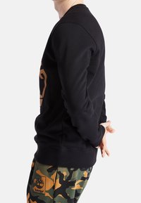 Sudadera negra con cuello redondo y puños de canalé, que presenta un gran logo dorado en la parte frontal, combinada con pantalones de camuflaje.