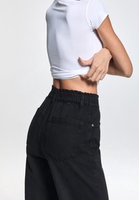 Svarta denimbyxor med en rynkad midja och sidofickor. Modellen bär en åtsittande vit kortärmad topp som avsl öjar midjan.