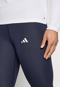 Mørkeblå tights med teksturert finish og reflekterende hvit Adidas-logo. En tettsittende hvit langermet topp med ribbet tekstur på kanten.