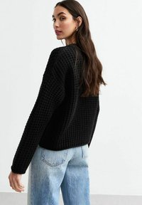 Zwart, getextureerd gebreid vest met een losse pasvorm en lange mouwen, gecombineerd met lichtblauwe denim jeans. De stof heeft een geweven patroon.