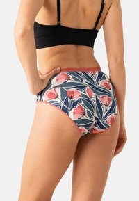 Femme portant des sous-vêtements à taille haute à motif floral avec des feuilles roses et bleues, associée à un débardeur court noir, la main sur la hanche.