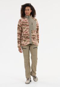 Veste en polaire avec un motif camouflage rose et vert, fermeture éclair à l'avant, avec une poche sur la poitrine, associée à un pantalon cargo beige et des chaussures de randonnée.