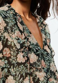 Blusa nera trasparente con un motivo floreale in rosa e verde. Presenta un collo a V e un design arricciato lungo la parte anteriore, realizzata in un tessuto leggero.