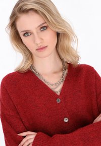 Rote Wolljacke mit V-Ausschnitt, zwei Knöpfen und langen Ärmeln. Akzentuiert durch eine mehrlagige silberne Kettenhalskette.
