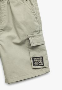 Shorts cargo color caqui claro, hechos de una tela suave, con un bolsillo lateral y una etiqueta negra con texto blanco. Bordes desgastados y costuras cosidas.