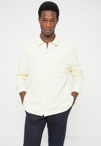 Ανοιχτό κίτρινο πουκάμισο με κουμπιά, γιακά και μια τσέπη στο στήθος, με μακριά μανίκια και υφασμάτινη υφή, σε συνδυασμό με σκούρο παντελόνι.