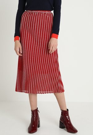 Kobieta ubrana w plisowaną spódnicę midi w kolorze czerwonym i srebrnym, czarną bluzkę z długim rękawem z czerwonymi mankietami oraz błyszczące bordowe botki za kostkę.