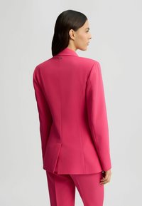 Blazer ajusté rose vif avec un revers cranté, une coupe cintrée et une fente arrière simple ; présente un logo discret au niveau du col.
