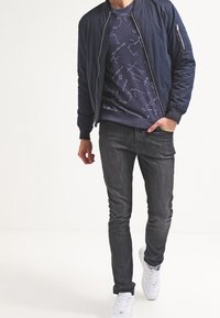 Giacca bomber blu navy con polsini a coste, una tasca con zip e una t-shirt nera con grafiche che presentano motivi delle costellazioni. Jeans scuri slim-fit e sneakers bianche.