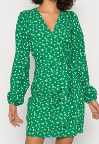 Vestido verde floral con escote en V y largas mangas abullonadas. Presenta un cinturón atado en la cintura y una tela texturizada con pequeñas flores blancas.